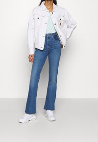 Chaqueta de mezclilla blanca con doble bolsillo, camiseta azul clara, jeans de tiro alto acampanados azules y zapatillas blancas. Acentos de hardware mínimos.