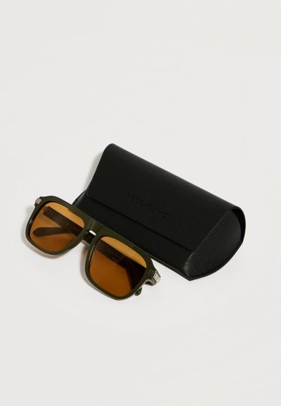 Ορθογώνια πράσινα γυαλιά ηλίου με κεχριμπαρένιους φακούς, τοποθετημένα δίπλα σε μαύρη σκληρή θήκη με υφή, πάνω σε λευκή επιφάνεια.