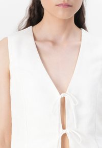 Witte linnen top met een diepe V-hals, een striksluiting in het midden en een minimalistische structuur zonder mouwen.