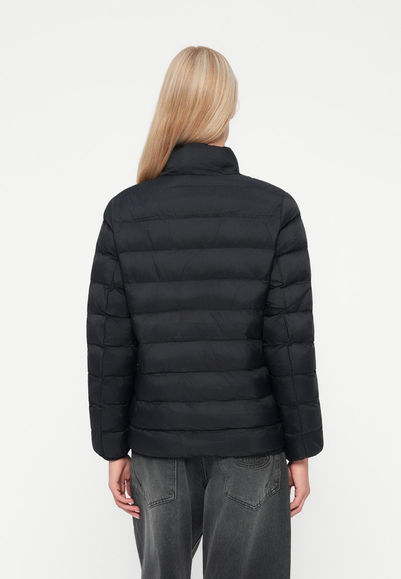 Chaqueta de plumas negra con patrón acolchado, cuello alto y mangas largas. Material suave y ligero. Combinada con jeans grises de corte relajado.