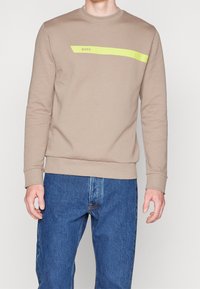 Beige sweatshirt med rund halsringning, med ett horisontellt neon gult streck med texten "BOSS". Kombineras med blå blåjeans.