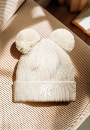 Gorro de punto color crema con dos pompones esponjosos y un logo bordado en blanco de los New York Yankees, colocado sobre una superficie de tela a rayas.