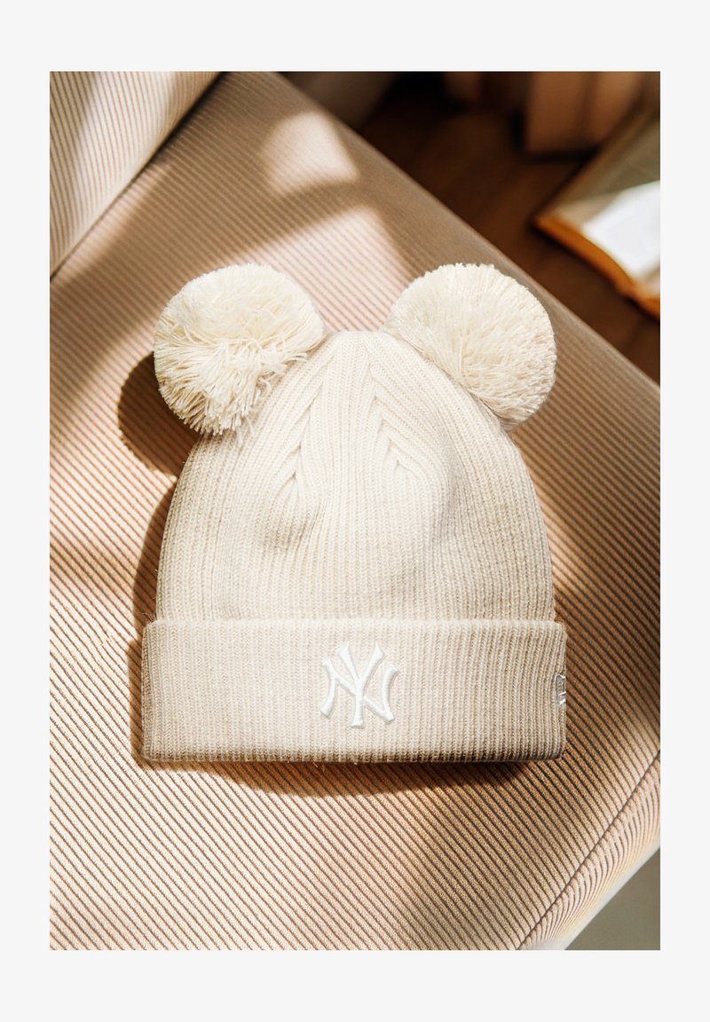 Kermanvärinen neulottu pipo, jossa on kaksi pörröistä pom-pomia ja valkoinen brodeerattu New York Yankees -logo, sijoitettu raidallisen kankaan päälle.