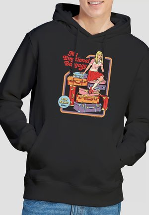 Lächende Person trägt einen schwarzen Hoodie mit buntem Motiv eines Mädchens, das auf aufgetürmten Koffern sitzt, beschriftet mit „My Emotional Baggage“ und „Life Skills Series“.