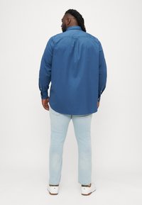 Modrá košeľa s dlhým rukávom a klasickým golierom; vyrobená z hladkej tkaniny. Spárovaná s svetlomodrými džínsami a bielymi teniskami. Detaily zahŕňajú gombíkové manžety.