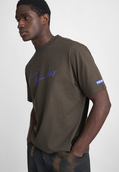 T-shirt in cotone marrone con testo scritto a mano blu "Blue Monday" sul davanti e un'etichetta blu sulla manica. Maniche corte e vestibilità rilassata.