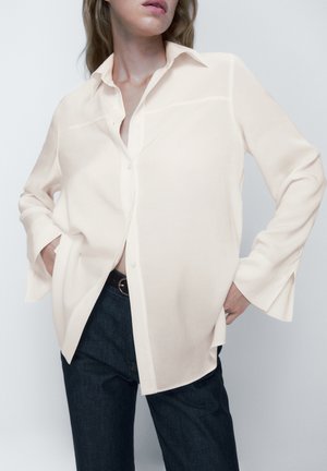 Woman wearing loose, cream-colored button-up shirt with hands in pockets, paired with dark jeans and a black belt, against a plain background.