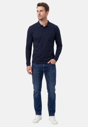 Uomo in piedi con la mano in tasca, indossa una polo a maniche lunghe blu navy, jeans blu e scarpe da ginnastica bianche su uno sfondo semplice.