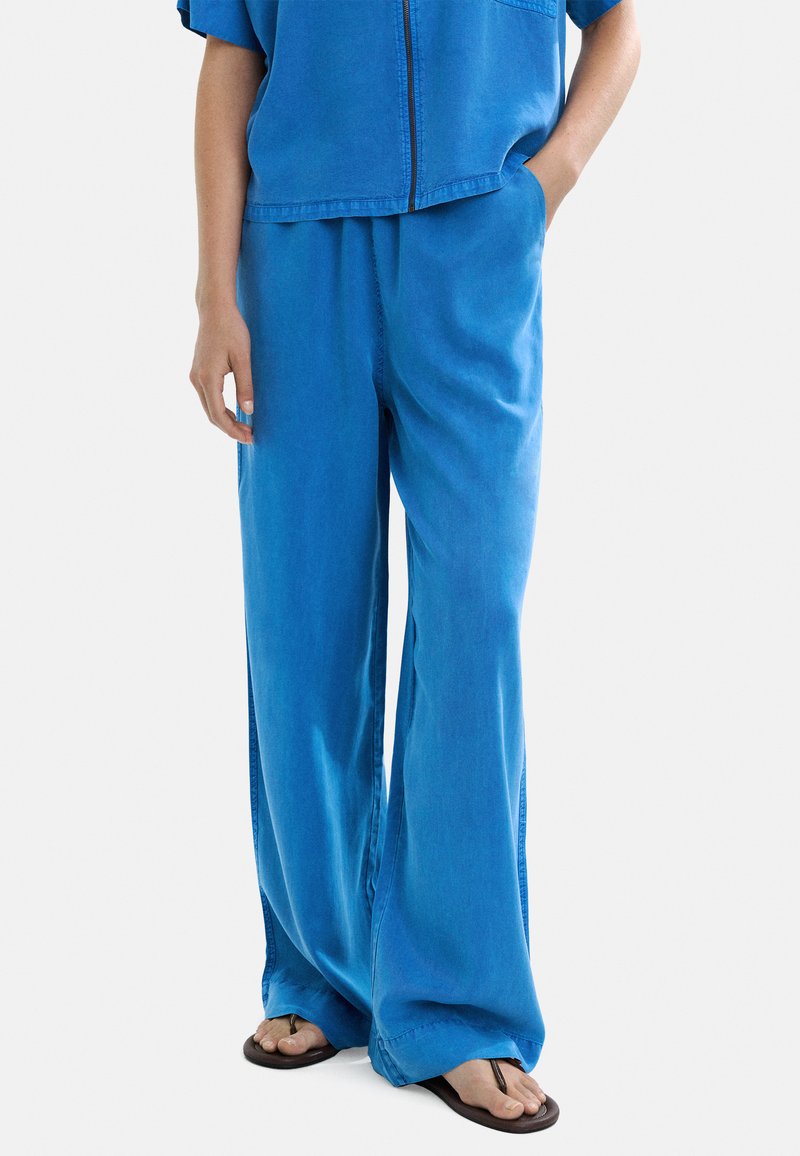 Osoba w luźnych niebieskich spodniach i pasującej bluzie na zamek, stojąca z jedną ręką w kieszeni i brązowych klapkach na stopach.