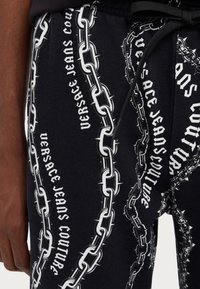 Črne trenirke z verižnim vzorcem v beli barvi in napisom "Versace Jeans Couture." Material izgleda mehak z teksturiranim videzom.
