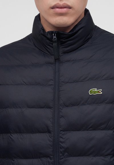 Homme portant une doudoune noire zippée jusqu'en haut avec un petit logo crocodile vert sur le côté gauche de la poitrine.