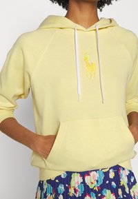 Gul huvtröja i mjukt material, med en framficka, vita snoddar och en liten broderad logotyp. Kombineras med en blommig kjol.