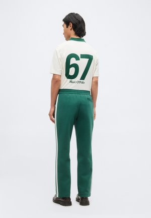 Homme portant un polo blanc avec un grand "67" vert et le texte Marc O'Polo, associé à un pantalon de survêtement vert avec des bandes blanches sur les côtés.