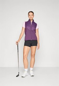 Γυναίκα που στέκεται με μια ρακέτα γκολφ, φορώντας ένα μωβ αμάνικο γιλέκο με φερμουάρ, ροζ κοντομάνικο μπλουζάκι, μαύρα σορτς, άσπρες κάλτσες και άσπρα αθλητικά παπούτσια.