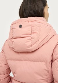 Lyserød puffjakke med høj krave og et sort logo detalje på ryggen. Materialet ser blødt og quiltet ud for isolering.