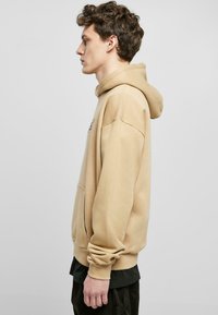 Beige kapucnis pulóver, puha anyag, laza szabás, kenguru zseb, bordázott mandzsetták és alsó szegély. A modell oldalra néz egy sima háttér előtt.