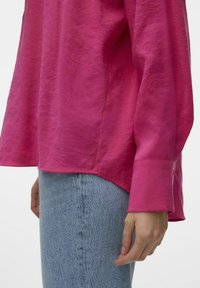 Fuchsia langærmet skjorte med en afslappet pasform og en subtil glans. Har en høj-lav kant og cuffs detaljer, parret med lyseblå denim.