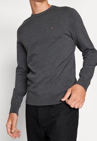 Grå crewnecktröja i stickad tyg. Har ribbade ärmslut och nederdel, med en liten logotyp på bröstet. Kombination med svarta byxor.