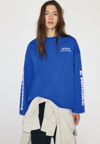 Sweat-shirt bleu à manches longues avec texte blanc indiquant "Come dalla Nonna TRATTORIA" et "AMORE" sur les manches. Coupe décontractée, matériau en coton.