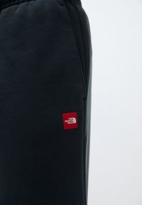 Črne trenirke iz mehkega materiala, z elastičnim pasom in majhnim rdečim logotipom na levi stegn.