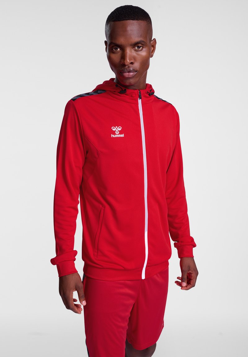 Rote zip-up Jacke mit Kapuze, aus glattem Material. Verfügt über einen weißen Reißverschluss, kontrastierende schwarze Schulterstreifen und Kängurutaschen.