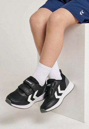 Barn iført sorte og hvide Hummel-sneakers med dobbelte velcrobånd, hvide sokker og blå shorts, siddende på en hvid blok.