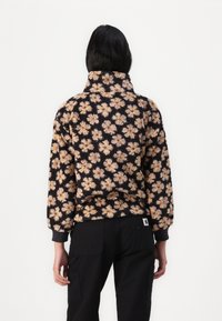 Sudadera de forro polar con fondo negro y patrón floral en melocotón. Cuello alto, corte holgado y puños elásticos. Pantalones negros con una etiqueta de logo visible.