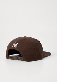 Brun baseball cap med en hvit brodert logo, flat skjerm og justerbar stropp bak. Laget av slitesterkt stoff.