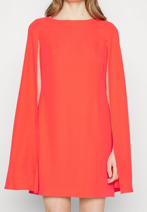 Femme portant une robe rouge vif avec de longues manches fendues ouvertes dévoilant des parties des bras, debout sur un fond blanc.