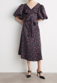 Robe portefeuille noire avec manches bouffantes et motif floral rose. Dotée d'une ceinture nouée à la taille et d'une jupe fluide, accompagnée de mules à talons noires.