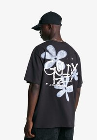 Schwarzes oversized T-Shirt mit blauen Blumenmotiven und weißem Text auf der Rückseite. Hergestellt aus Baumwolle, mit lockerem Schnitt und kurzen Ärmeln.