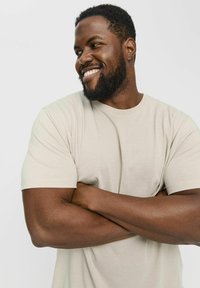 T-shirt beige à manches courtes en tissu doux, avec un design classique à col rond et une coupe décontractée, porté par une personne aux bras croisés.
