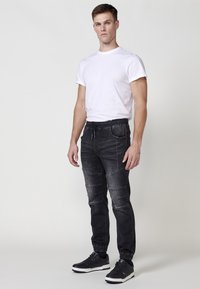 Pantalones joggers de mezclilla negra con cintura y puños elásticos, con paneles texturizados y bolsillos frontales, combinados con una camiseta blanca lisa y zapatillas negras.