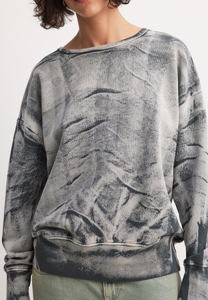 Frau trägt einen lockeren grau-schwarzen Batik-Sweatshirt mit langen Ärmeln und rundem Ausschnitt, kombiniert mit hellblauen Jeans.