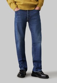 Jeans din denim albastru închis cu croială drept și o ușoară decolorare. Asortați cu pantofi negri cu șireturi și un pulover galben.