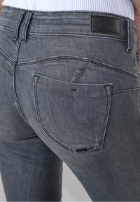 Detailní záběr na osobu v těsných tmavě šedých džínách, viditelná zadní kapsa a černý kožený štítek značky na pase.