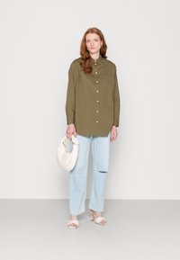 Camisa verde-oliva de botões com um bolso no peito, jeans rasgados de tom azul-claro, bolsa branca texturizada e sandálias de salto brancas.