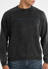 Zwarte pullover trui met een geribbelde ronde hals, lange mouwen en "GUESS ORIGINALS" geborduurd over de borst met sterren.