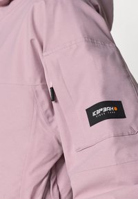 Világos rózsaszín dzseki, amely texturált anyagból készült, fekete márkajelzéssel a ujján, ami az "ICEPEAK since 1996" feliratot viseli.