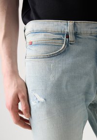 Šviesiai mėlyni džinsai su siauru siluetu, turintys mažą raudoną etiketę, pažeistą detalę ant kelio ir matomus siūlus.