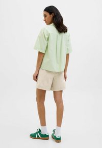 Camisa de manga corta a rayas verdes claras combinada con pantalones cortos beige. El modelo lleva zapatillas deportivas verdes con acentos blancos y calcetines blancos.