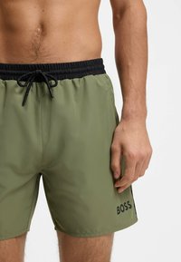 Ανδρικός κορμός που φοράει μαγιό σε ελαιώδες πράσινο με μαύρη ελαστική ζώνη μέσης και κορδόνι, με λογότυπο "BOSS" στο αριστερό πόδι.