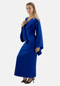 Długa niebieska sukienka z dekoltem w kształcie litery V, taliowana, z szerokimi rękawami. Wykonana z gładkiej tkaniny, sięga do podłogi, zestawiona z czarnymi szpilkami.