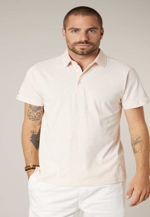 Homme aux cheveux courts et à la barbe, portant un polo rose clair et un pantalon blanc, montrant des tatouages sur les deux avant-bras et des bracelets au poignet gauche.