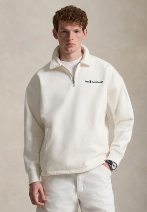 Mladý muž s kučeravými červenými vlasmi, ktorý má na sebe bielu polovične rozopnutú mikinu Polo Ralph Lauren a biele nohavice, stojaci pred neutrálnym pozadím.