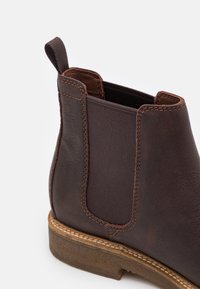 Bottine en cuir marron avec panneaux élastiques sur les côtés, détails cousus, bout arrondi et semelle extérieure texturée avec un talon discret.