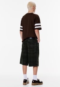 Bruine kortekorte mouwen shirt met witte strepen, gecombineerd met donkergroene geruite shorts. Zwarte schoenen en witte sokken maken de outfit compleet.