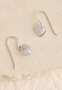 Silberne herzförmige Ohrringe mit polierter Oberfläche, versehen mit einem detaillierten Muschelrand, die an einem einfachen Haken-Design hängen.