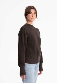 Brauner gerippter Strickpullover mit hohem Kragen und langen Ärmeln, der ein strukturiertes Muster und einen taillierten Bund aufweist, kombiniert mit hellblauen Jeans.
