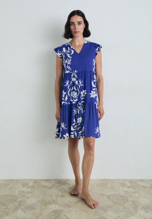 Abito blu con motivo floreale bianco, maniche corte, scollatura a V, design a strati. Tessuto leggero, che arriva a metà coscia, indossato scalzo.